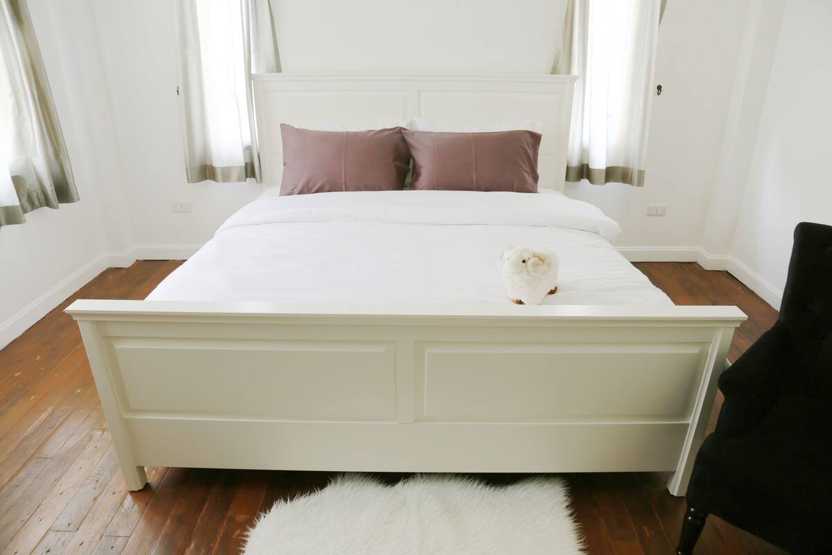 Uma cama branca com duas almofadas, um tapete branco e fofo, e uma cadeira preta.
