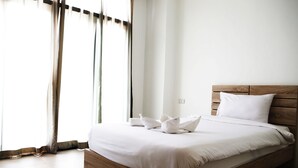 Un lit bien fait, avec du linge de lit blanc et des oreillers, une tête de lit en bois et une vue sur une fenêtre ornée de rideaux légers.