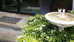 Une pancarte d’accueil au-dessus d’une porte vitrée, une grande plante en pot et un abreuvoir à oiseaux agrémenté de fleurs blanches.