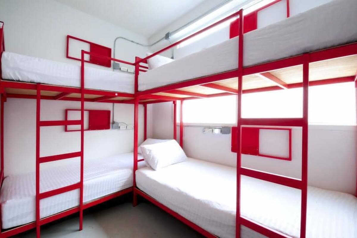 Een slaapkamer met stapelbedden, rode frames, witte beddengoed en een raam dat voor natuurlijk licht zorgt.