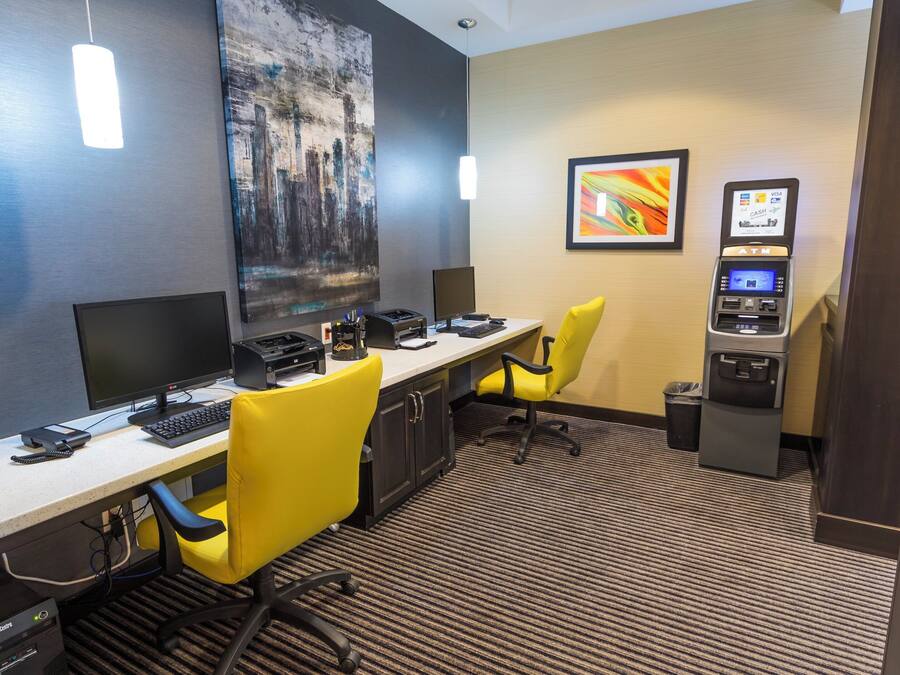 A hotel room with a desk, two chairs, a computer, an ATM, and a painting on the wall.