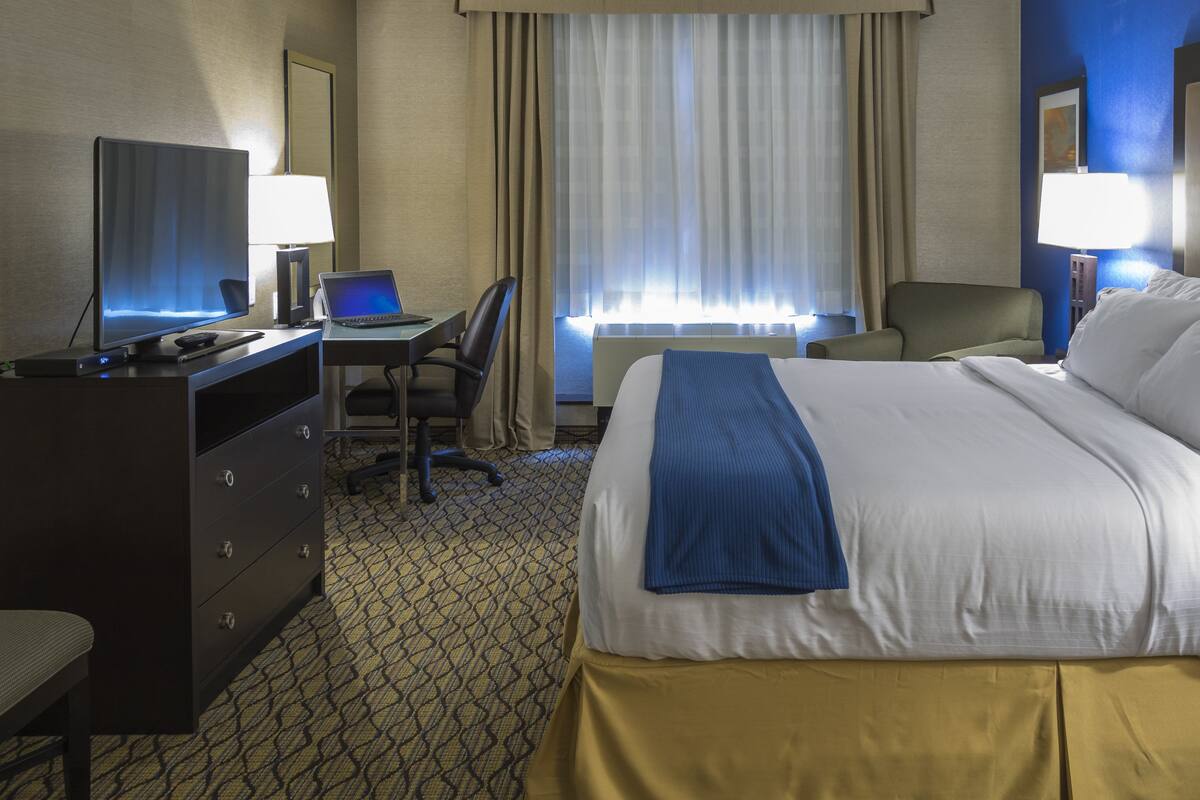 A hotel room with a bed, a desk with a laptop, a chair, a dresser, and a TV.