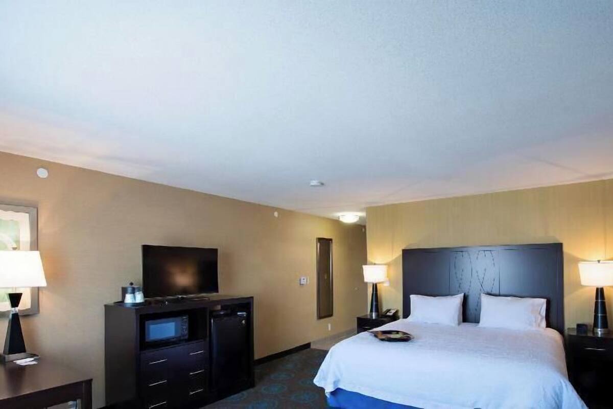 Habitación de hotel con cama, mesitas de noche, televisor, escritorio y ordenador, además de un cuadro en la pared.