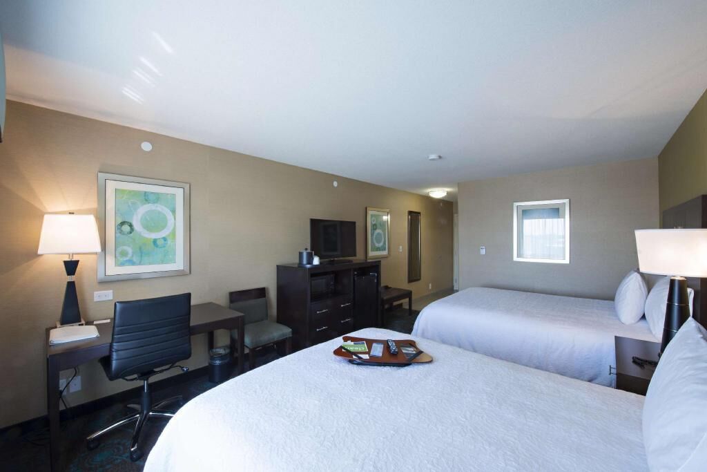 A hotel room with two beds, a desk, a chair, a TV, and artwork on the walls.