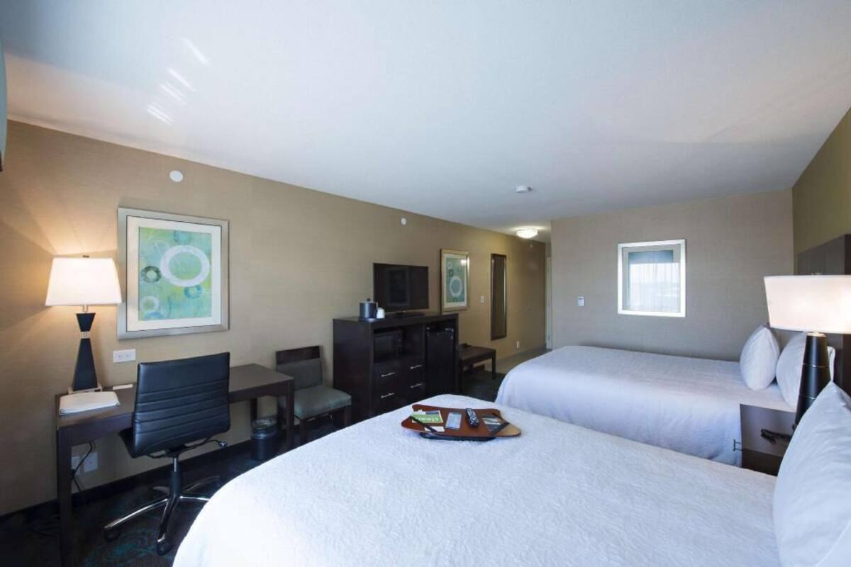 Habitación de hotel con dos camas, un escritorio, una silla, un televisor y obras de arte en las paredes.