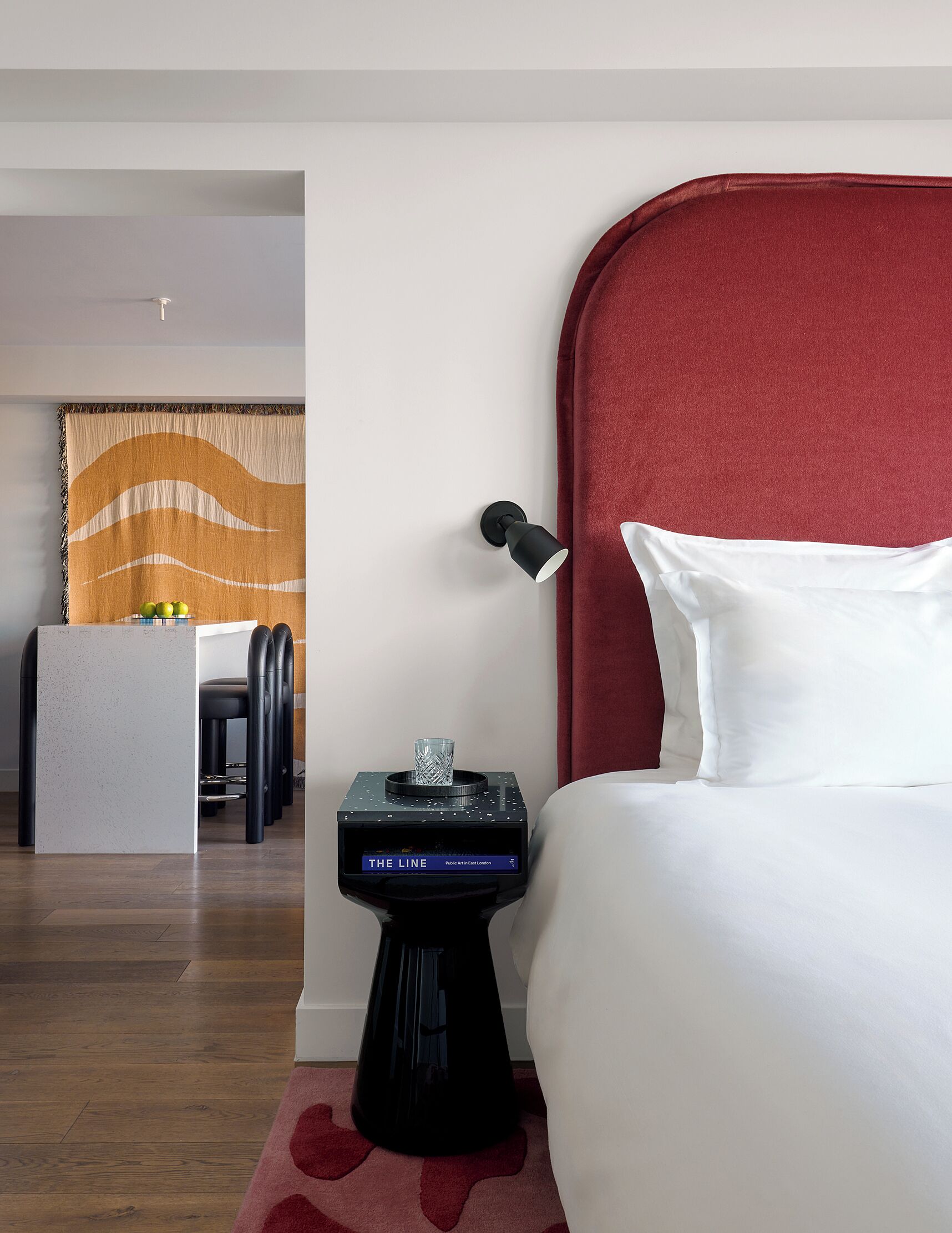 Une chambre d’hôtel avec une tête de lit rouge, une table de chevet avec une lampe, une petite table d’appoint ornée d’un objet décoratif, et une vue sur la salle à manger.