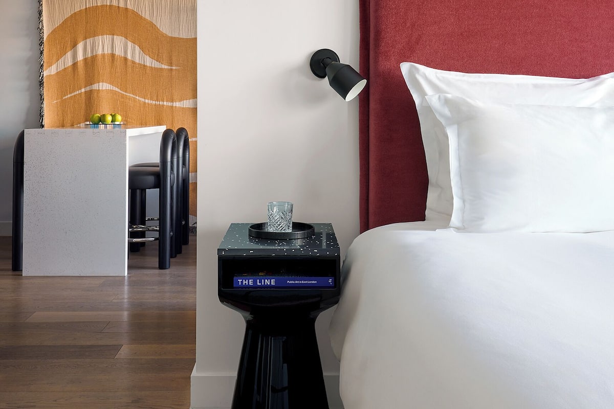 A hotel room with a red headboard, a bedside table with a lamp, a small side table with a decorative item, and a view into the dining area.