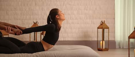 A person in a black top and leggings performing a stretching exercise on a white cushion in a room with a brick wall and two wooden lanterns.