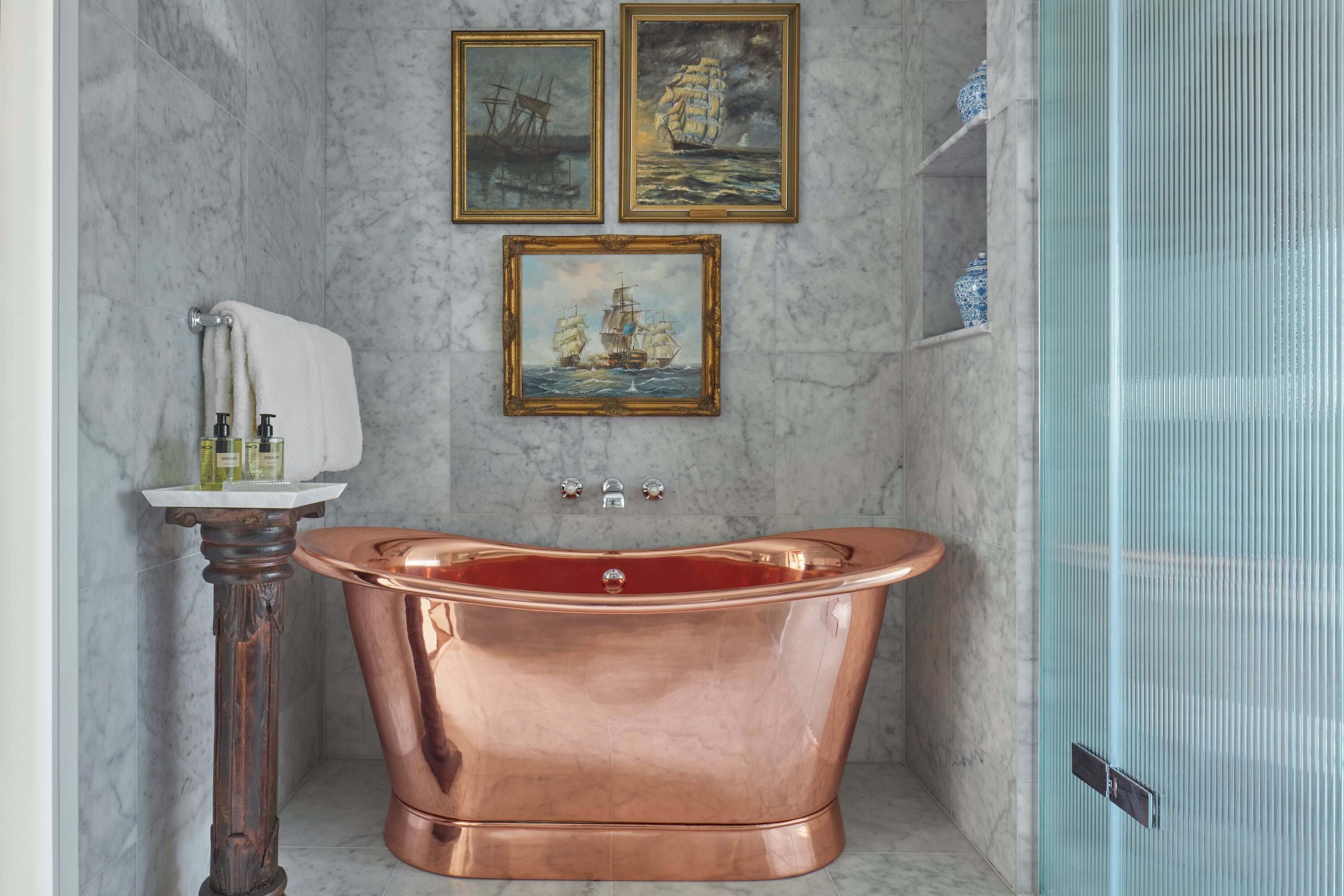 A bathroom with a copper bathtub, a pedestal sink, and two framed paintings on the wall.