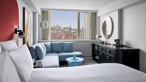 Habitació d'hotel moderna amb un llit gran, un sofà blau, una taula rodona petita, un prestatge negre i una finestra gran amb vista a la ciutat.