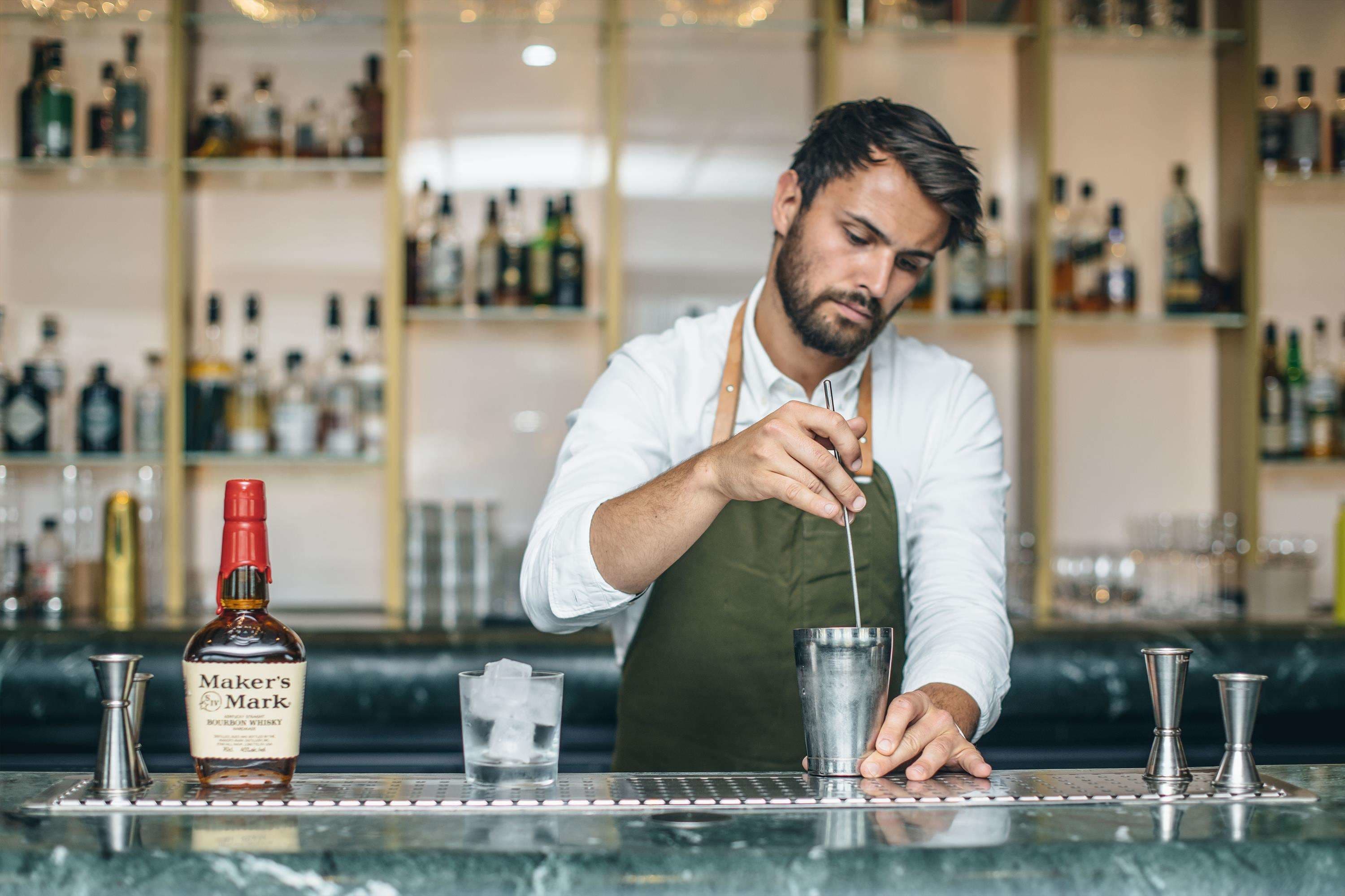 Een bartender bereidt een drankje toe aan de bar, met een fles Maker's Mark bourbon whisky en diverse baraccessoires.