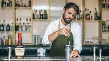 A bartender preparing a drink at a bar with a bottle of Maker's Mark bourbon whiskey and various bar tools.