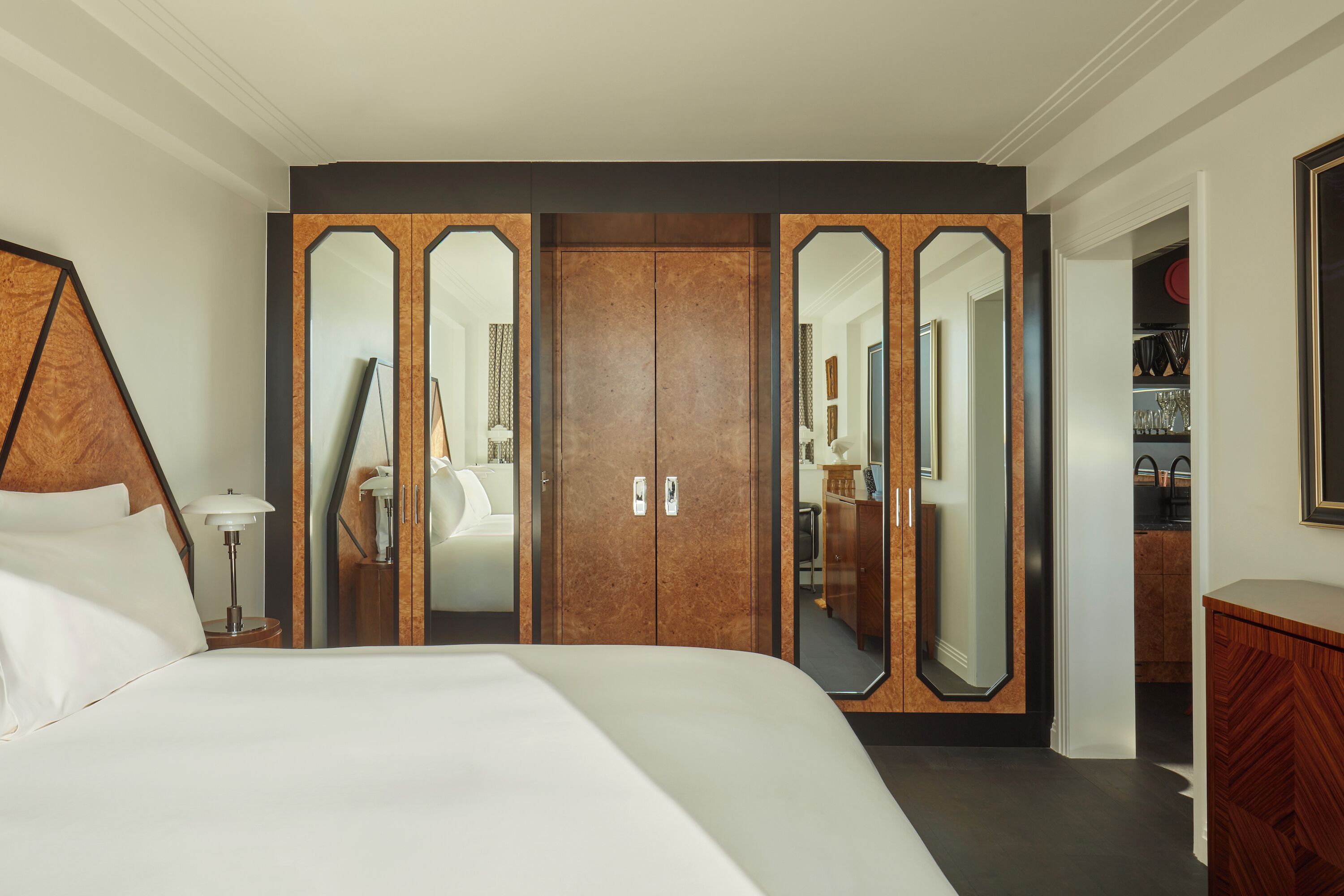 A hotel room with a large bed, a wardrobe with mirrored doors, a bedside table with a lamp, and a dresser.