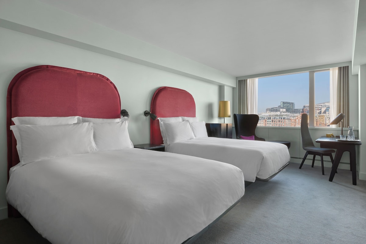 Ett hotellrum med två sängar, ett skrivbord, en stol och ett stort fönster med utsikt över staden.