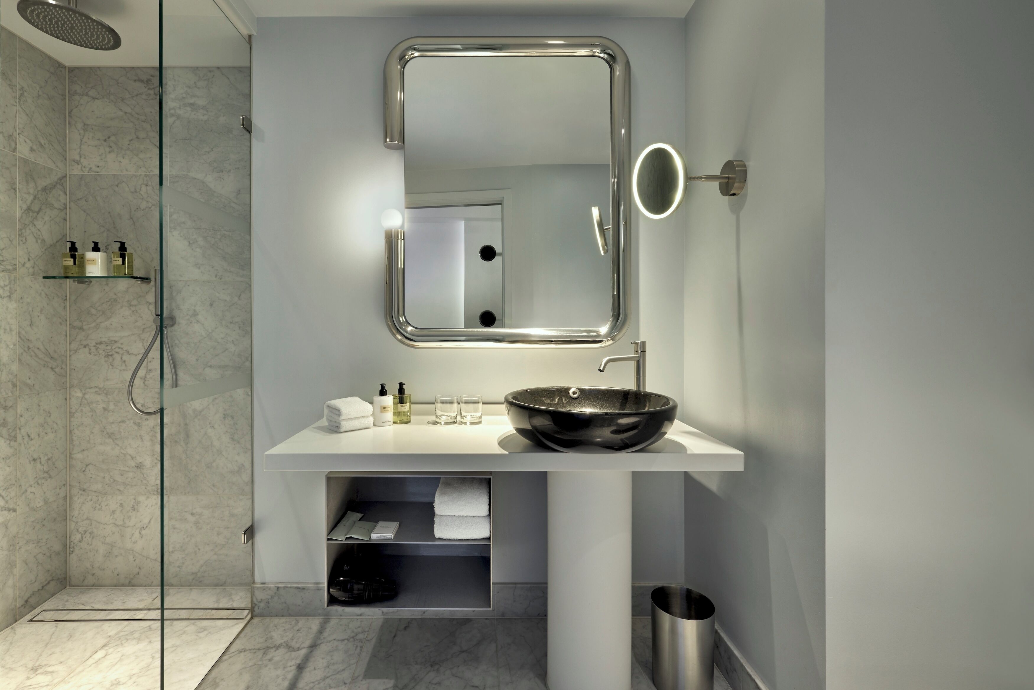 A modern bathroom with a glass-enclosed shower, a white sink with a black basin, a mirror with a light, and a small shelf with toiletries.