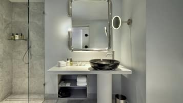 A modern bathroom with a glass-enclosed shower, a white sink with a black basin, a mirror with a light, and a small shelf with toiletries.