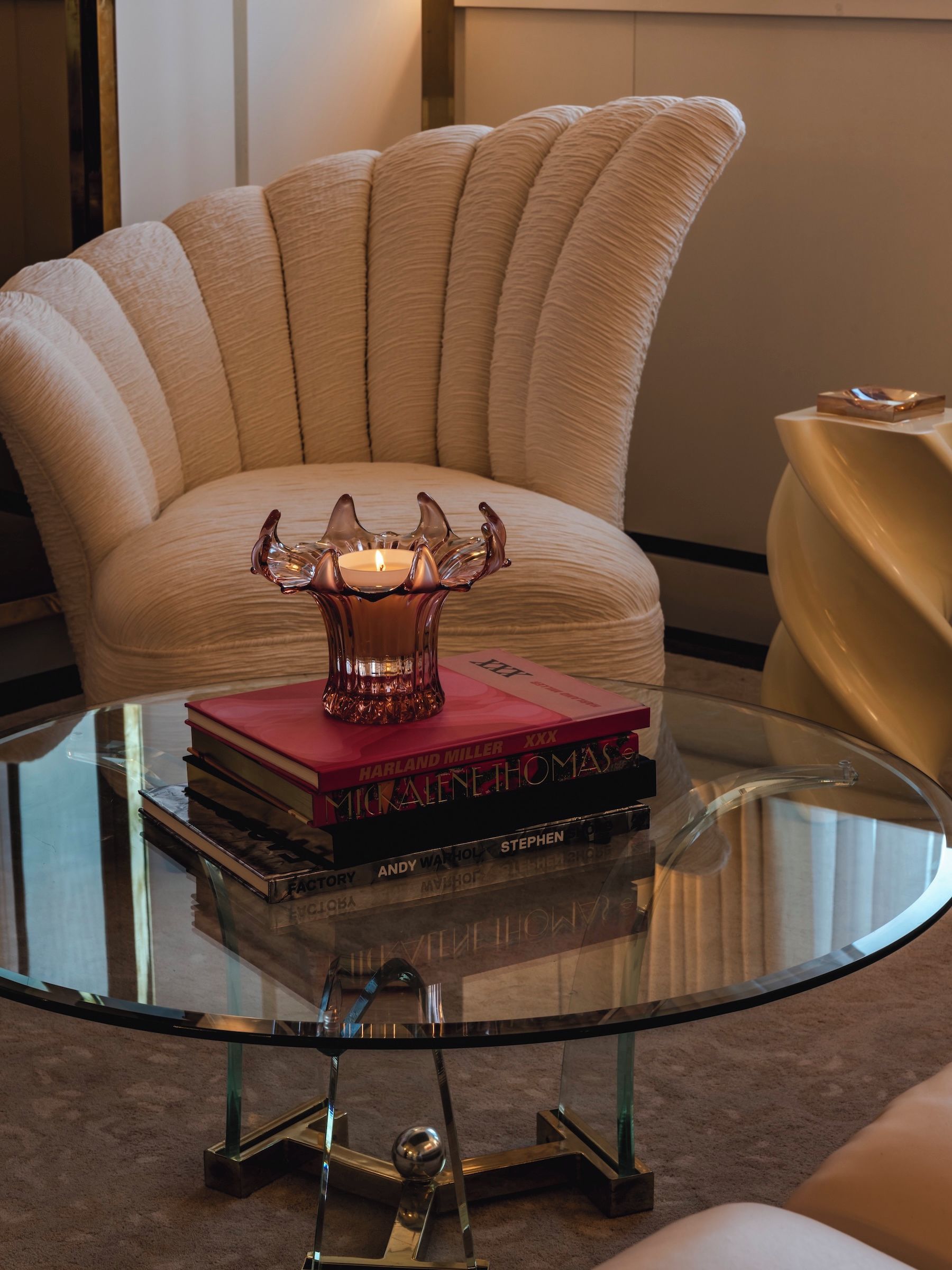 A glass coffee table with a candle in a glass holder, a stack of books, and a chair in the background.