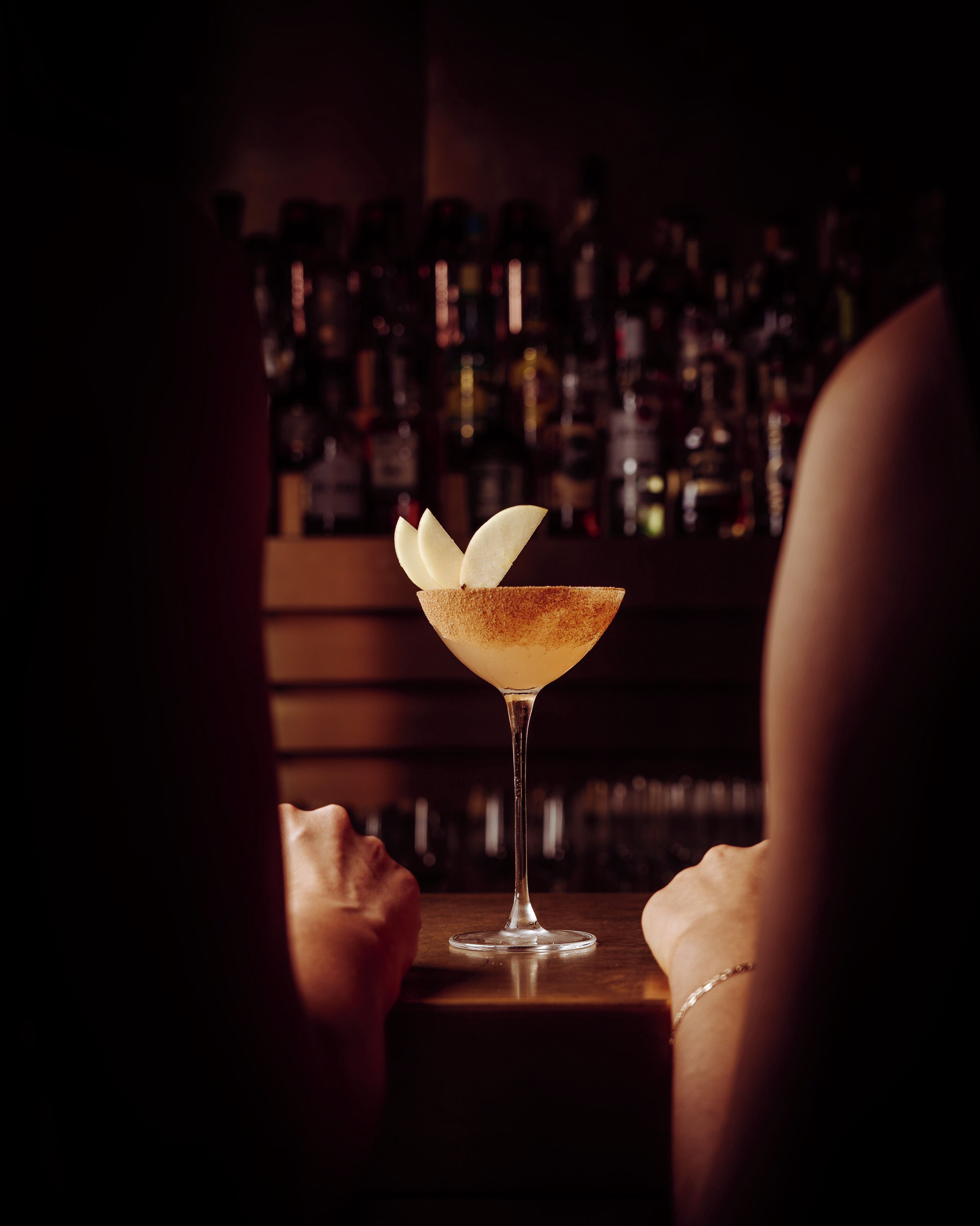 A bar setting with a martini glass on a table, a person partially visible, and a background of shelves with bottles.