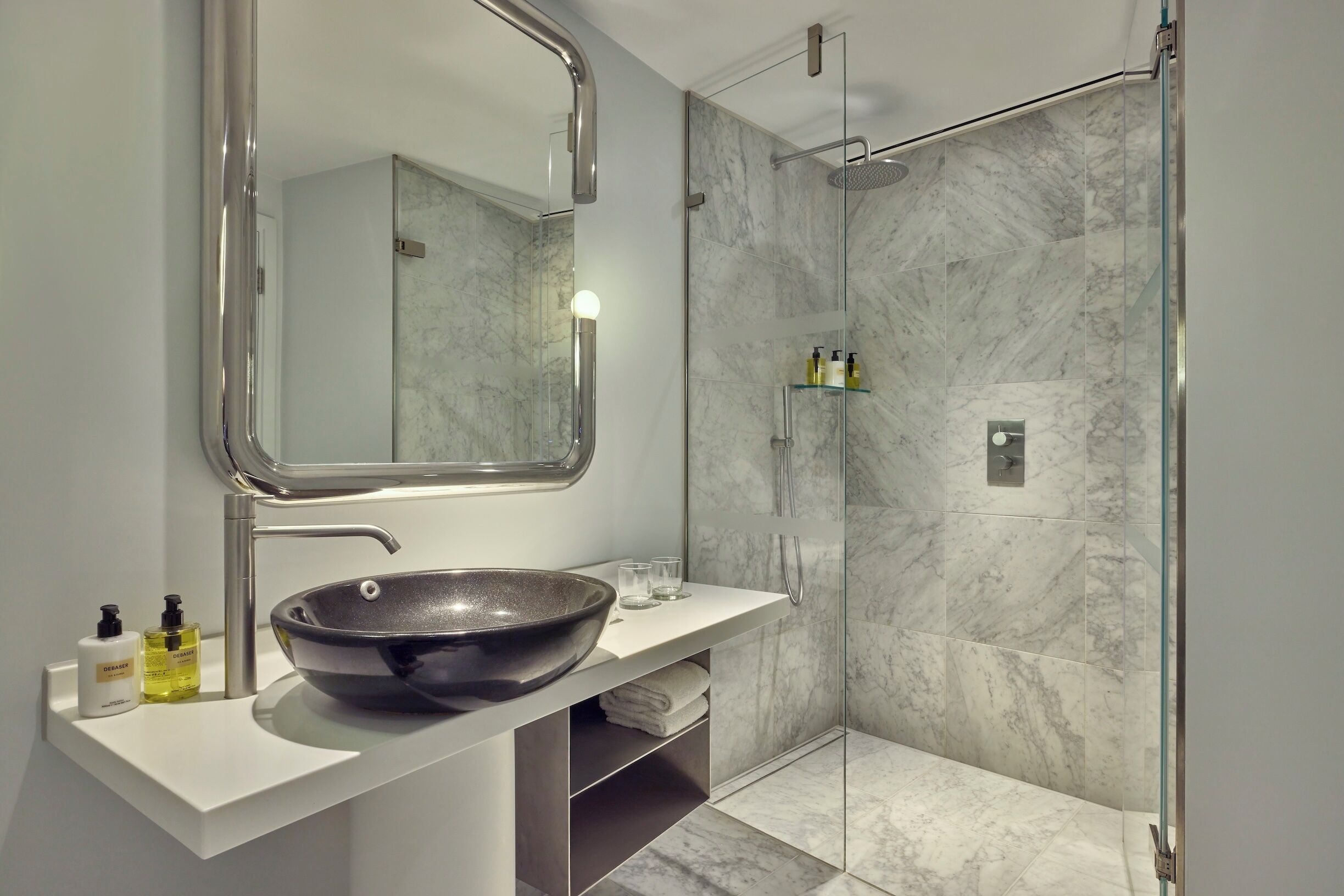A modern bathroom with a large mirror, a bowl sink, a glass shower enclosure, and marble tiles.