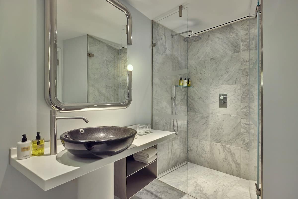A modern bathroom with a large mirror, a bowl sink, a glass shower enclosure, and marble tiles.