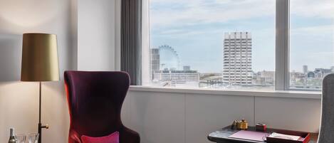 A modern hotel room with a large window offering a city view, a plush red chair, a small table with a lamp, and a desk with a tray of items.