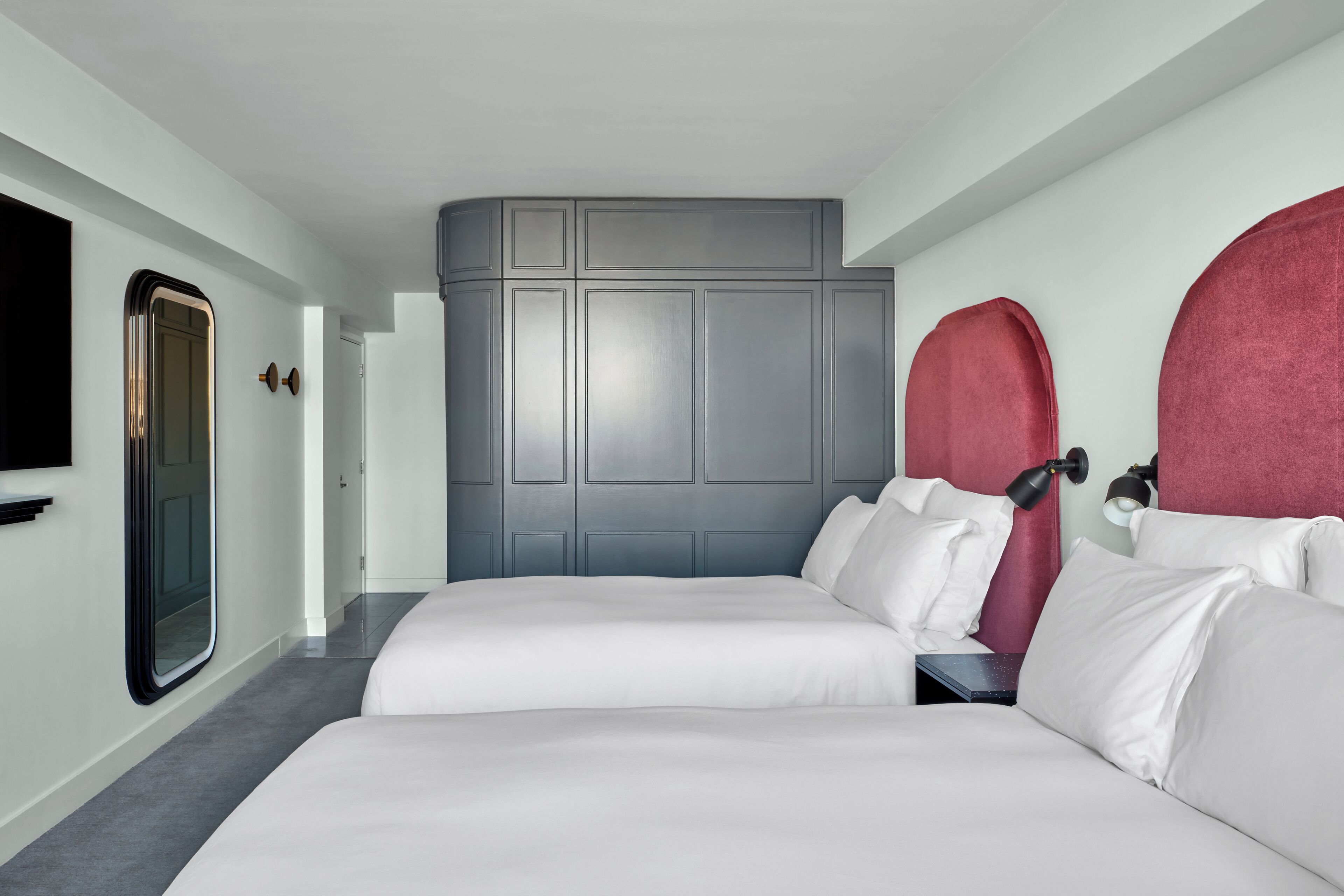 Une chambre d’hôtel avec deux lits, une tête de lit grise, un téléviseur et un mur d’accent rouge.