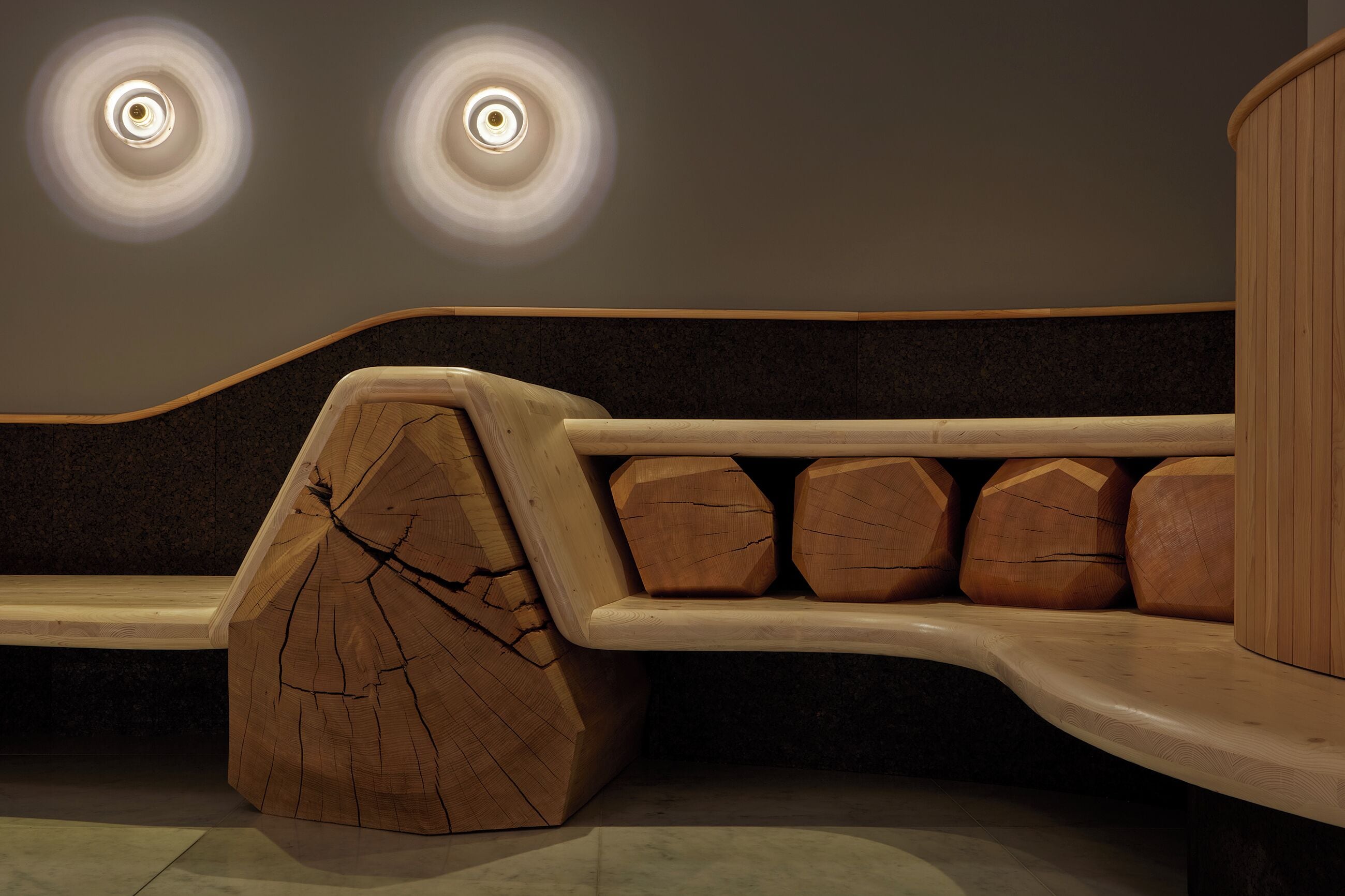 A wooden bench with a unique design, a wall with two circular light fixtures, and a curved wooden structure.