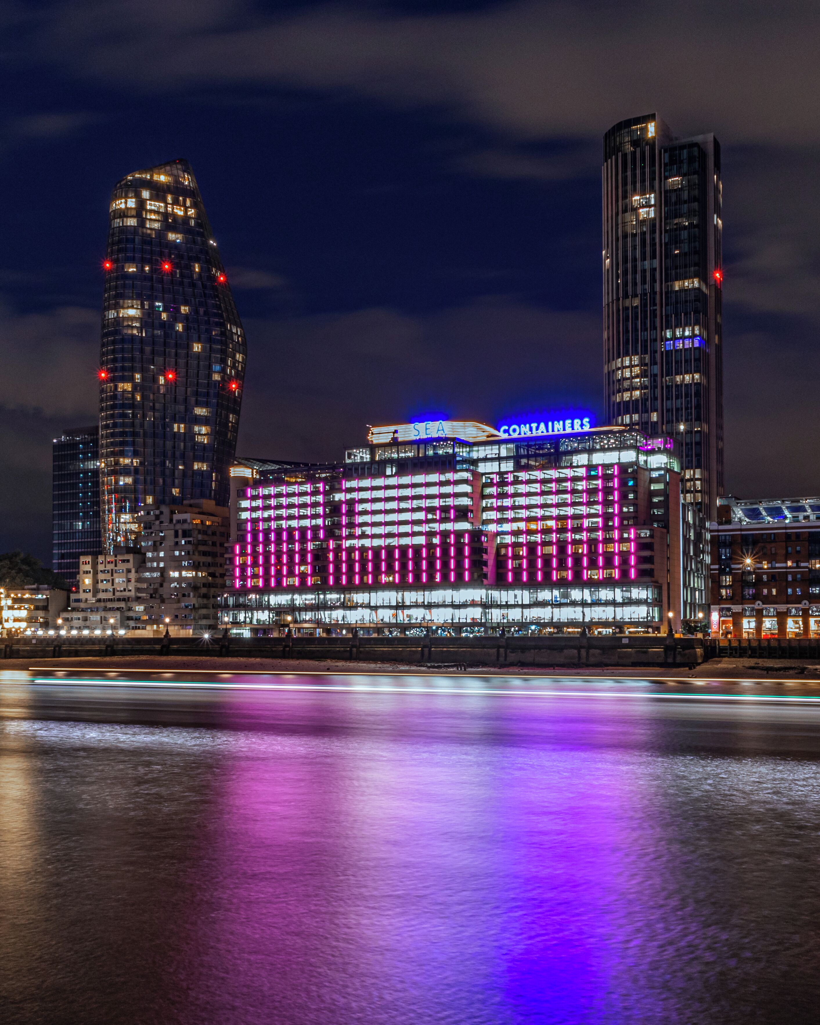 A waterfront hotel with illuminated signage, a prominent building with a curved facade, and a reflection on the water.