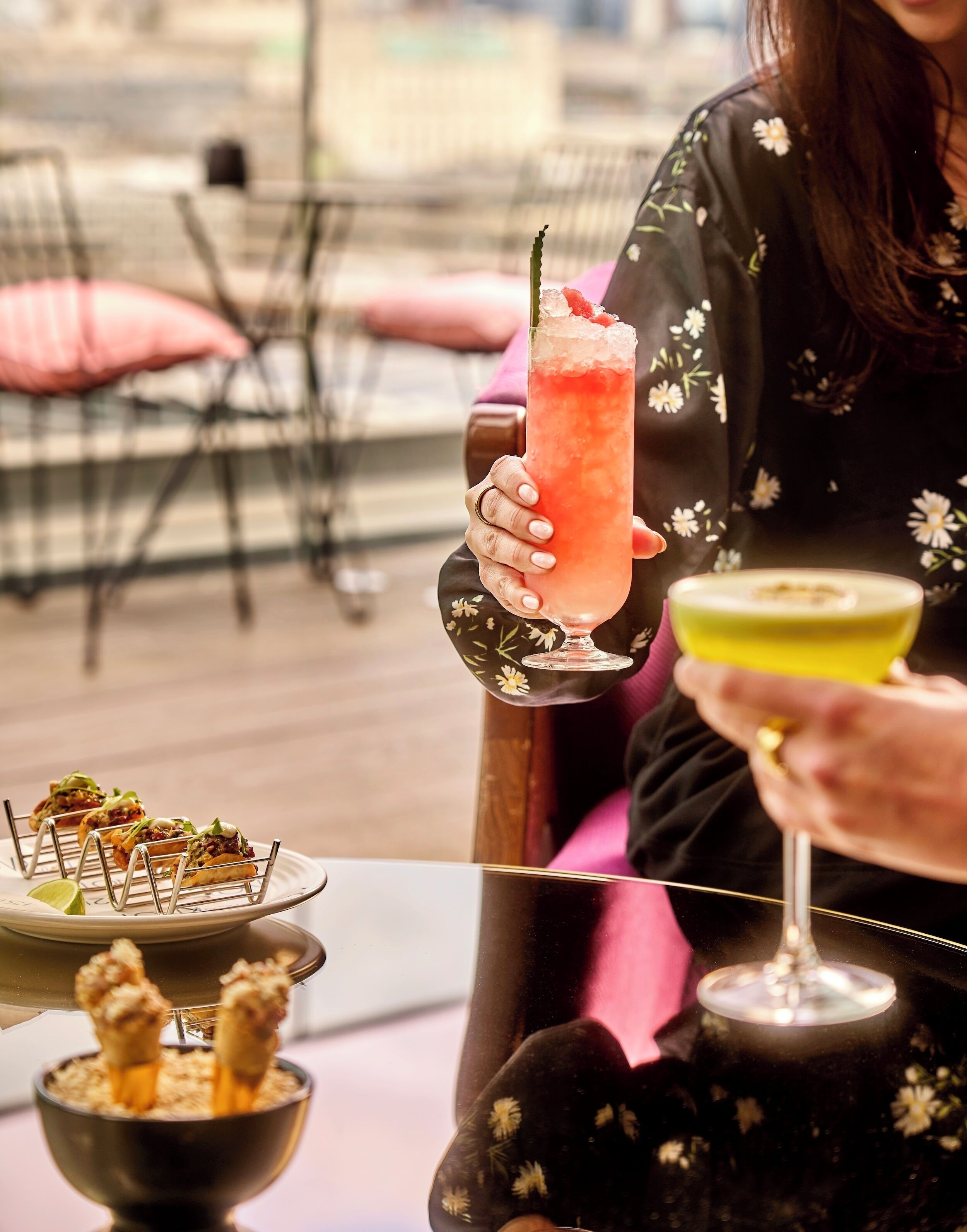 Eine Person hält einen rosa Cocktail mit einer Garnitur, einen gelben Cocktail und ein Teller mit Vorspeisen auf einem Tisch.