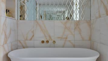 Una bañera independiente en un baño con paredes de mármol y un recinto con espejo.