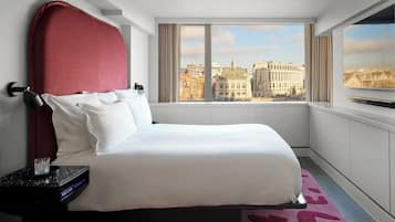 A modern hotel room with a large bed, a nightstand with a clock, a TV mounted on the wall, and a city view through the window.
