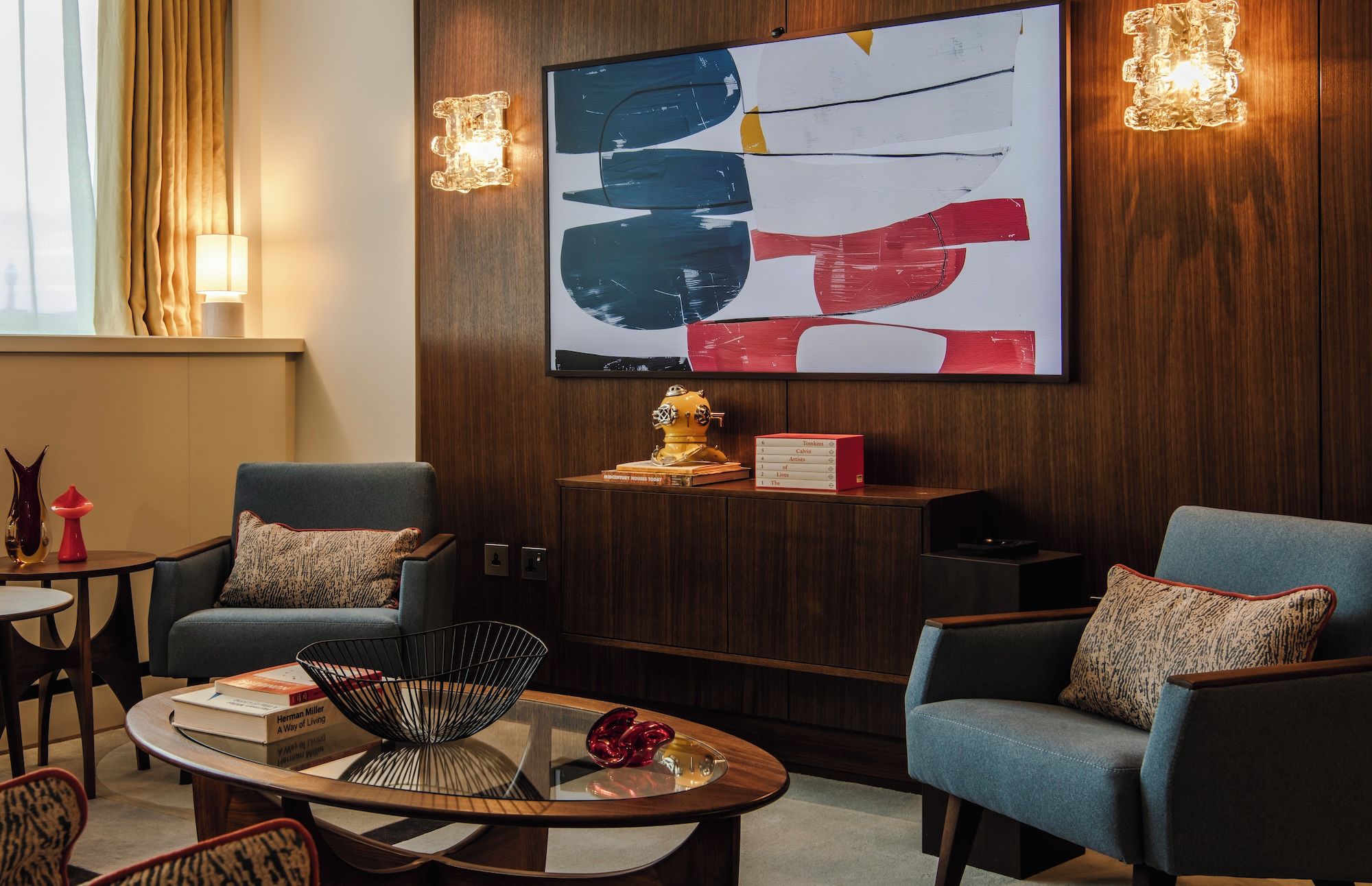 A modern hotel room with a large abstract painting, two armchairs, a small table with a book and a decorative bowl, and a wall-mounted lamp.