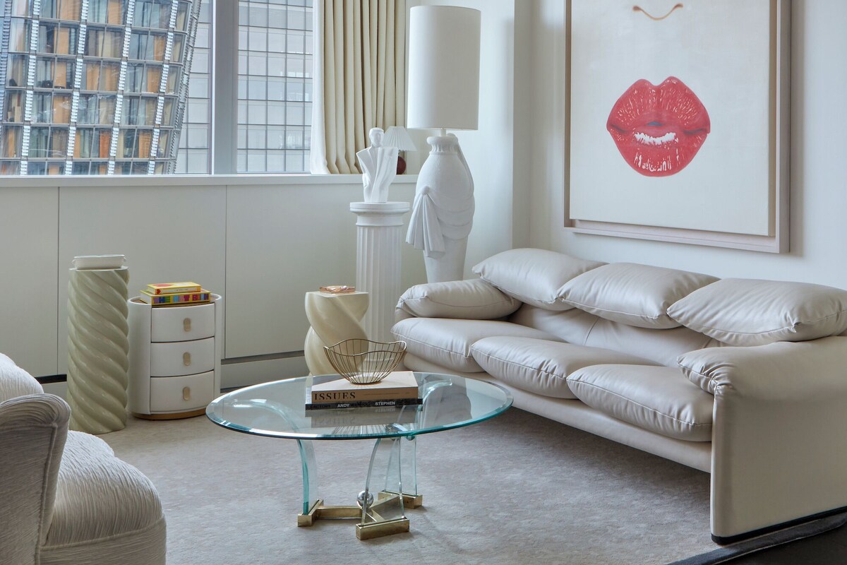 Un salon moderne avec un canapé blanc, une table basse en verre et une œuvre d’art encadrée représentant des lèvres.