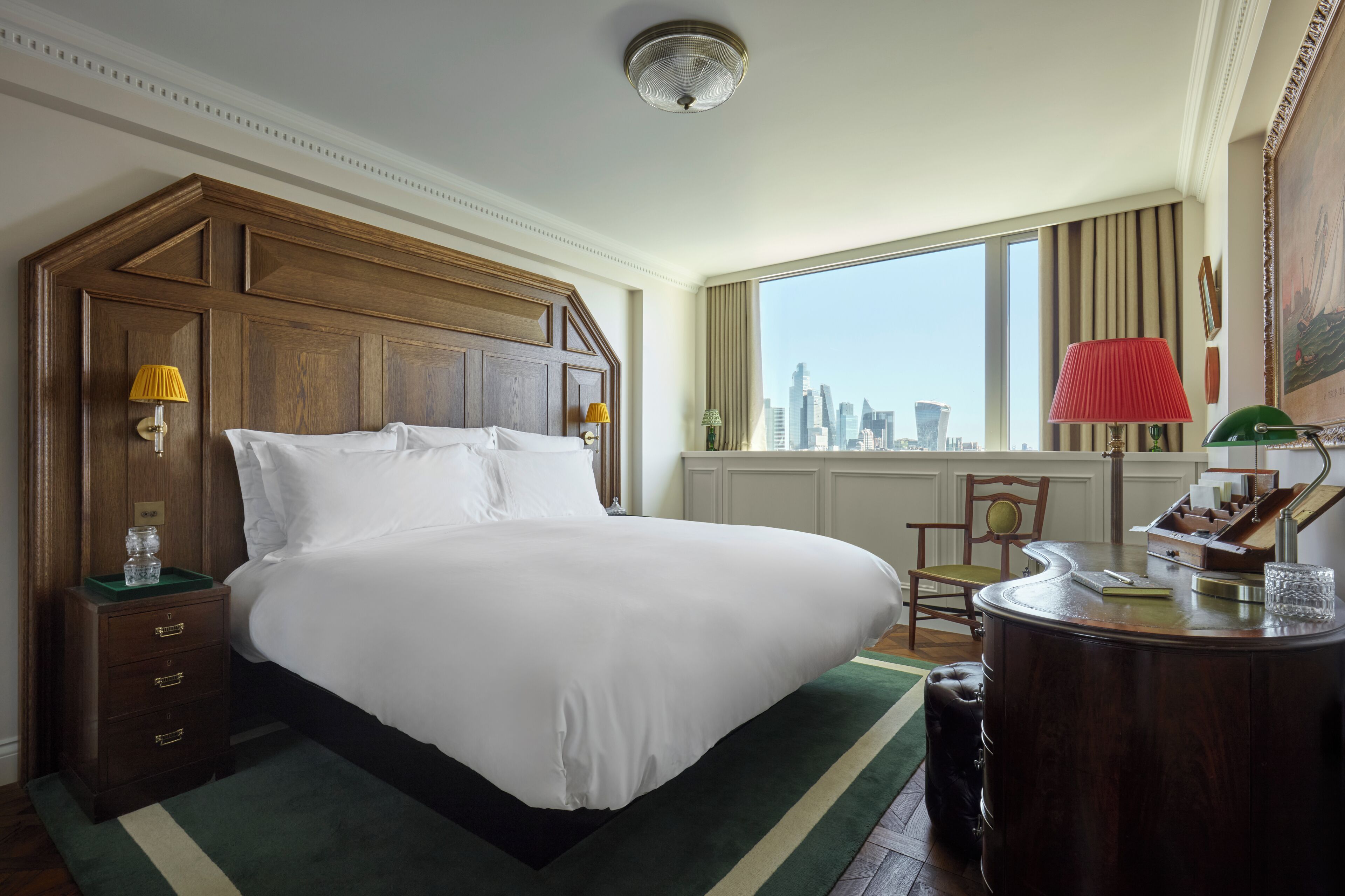 A large bed with a wooden headboard, a nightstand with a lamp, a desk with a red lamp, and a view of the city skyline through large windows.