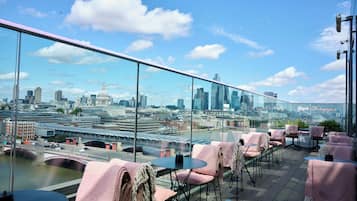 Eine Dachterrasse mit Tischen, Stühlen, einer Glastrennwand und Blick auf die Skyline der Stadt.