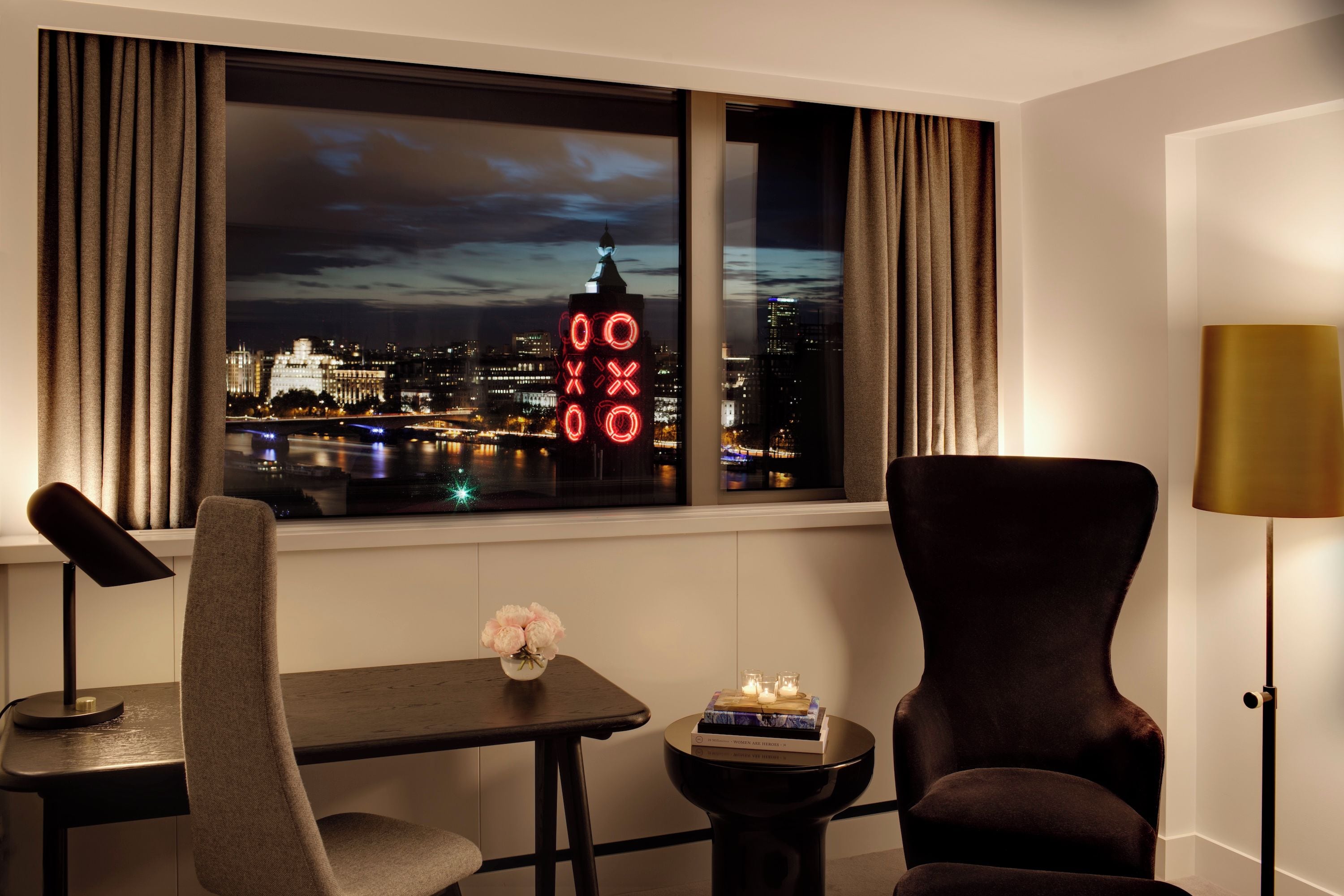 A hotel room with a view of a cityscape at night, a desk with a chair, a small table with a vase of flowers, and a lamp.