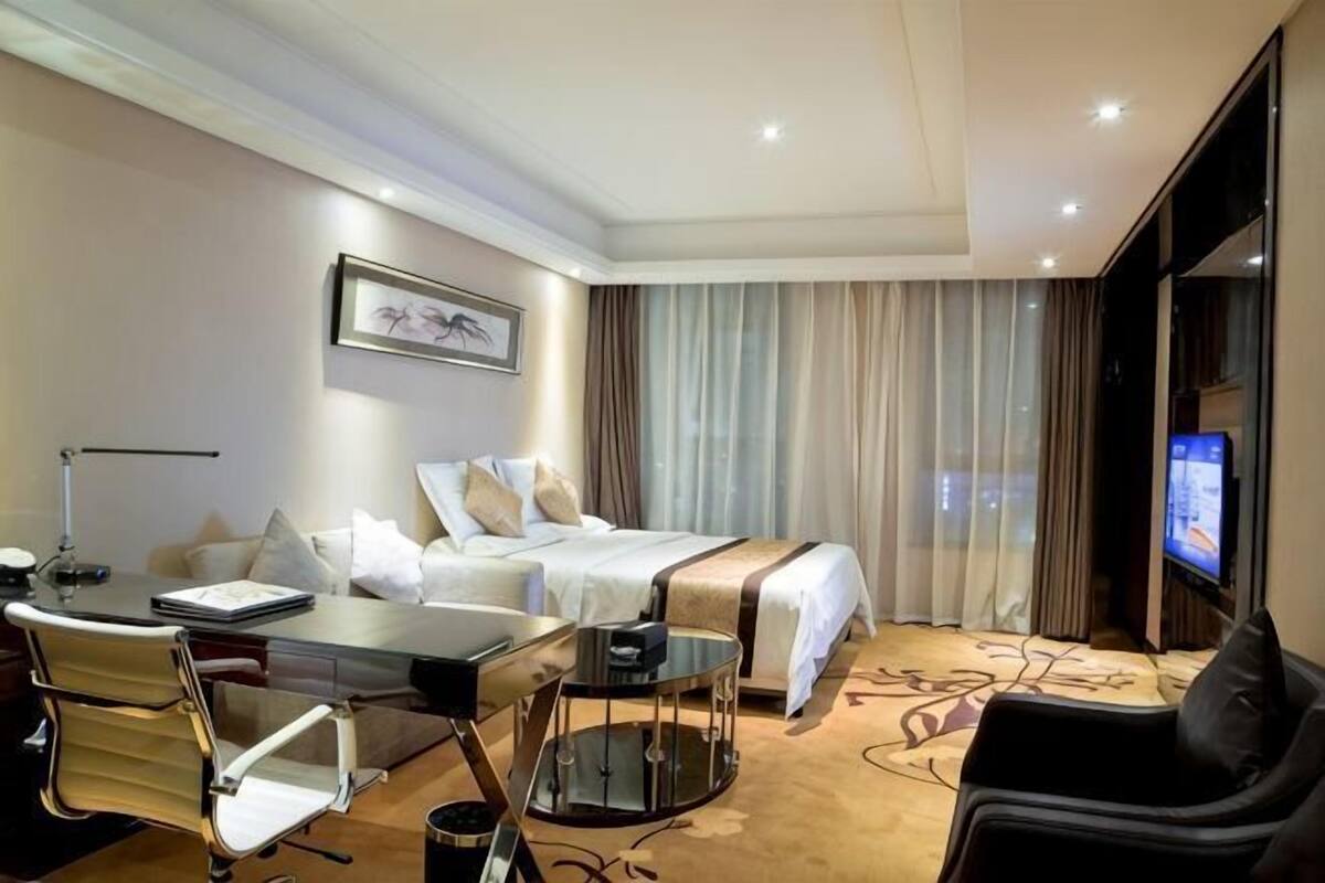 A hotel room with a bed, a desk with a chair, a TV, and a window with curtains.