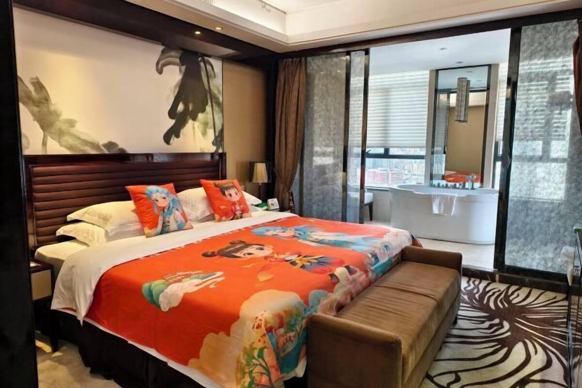 A hotel room with a large bed, a bench, a desk, a window with curtains, and a bathroom visible through the window.
