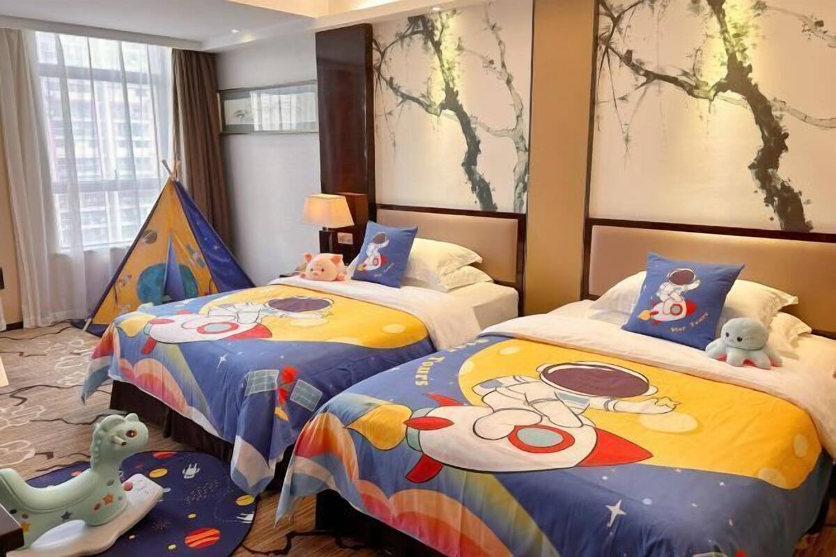 A hotel room with two beds, each with a colorful bedspread featuring cartoon characters. There is a large window with curtains, a painting on the wall, and a small table with a lamp.