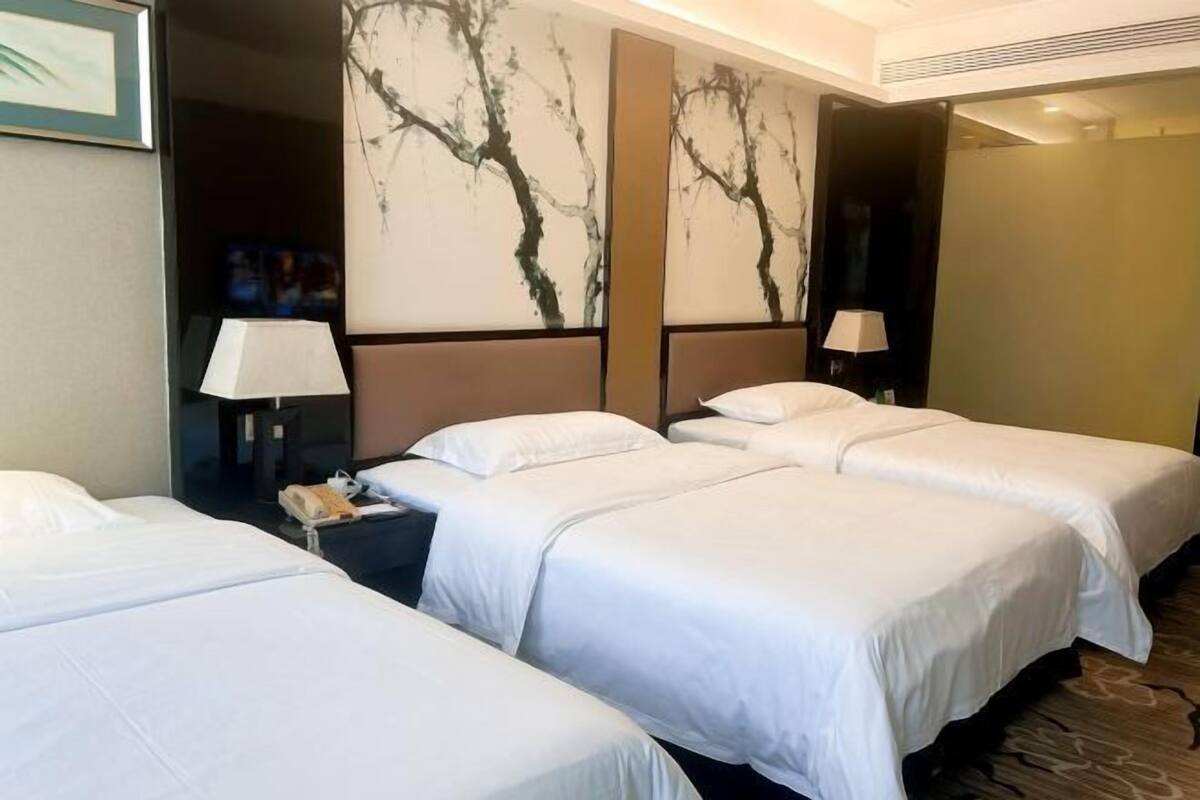 A hotel room with three beds, a nightstand with a lamp, and a wall with a tree painting.