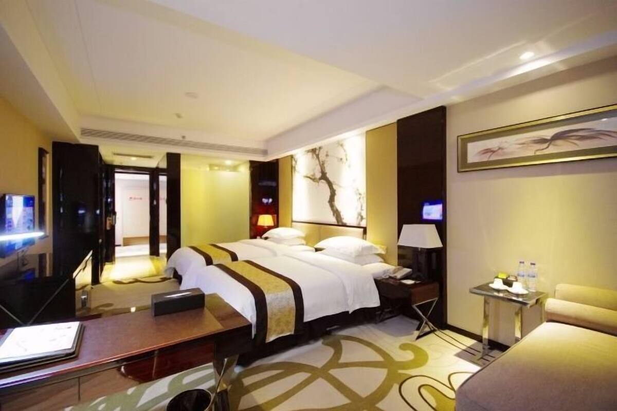 A hotel room with a large bed, a desk, a chair, a TV, and a small table with water bottles.