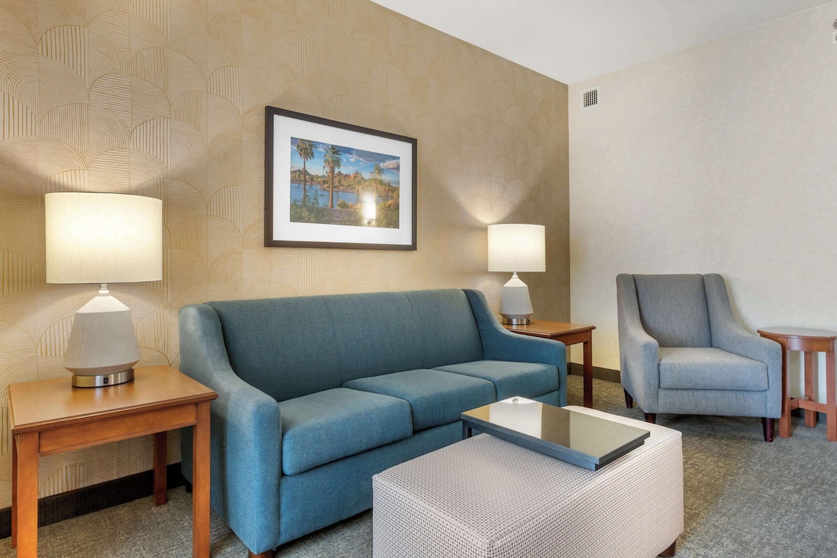 A hotel room with a blue sofa, a grey armchair, a small table, and two lamps.