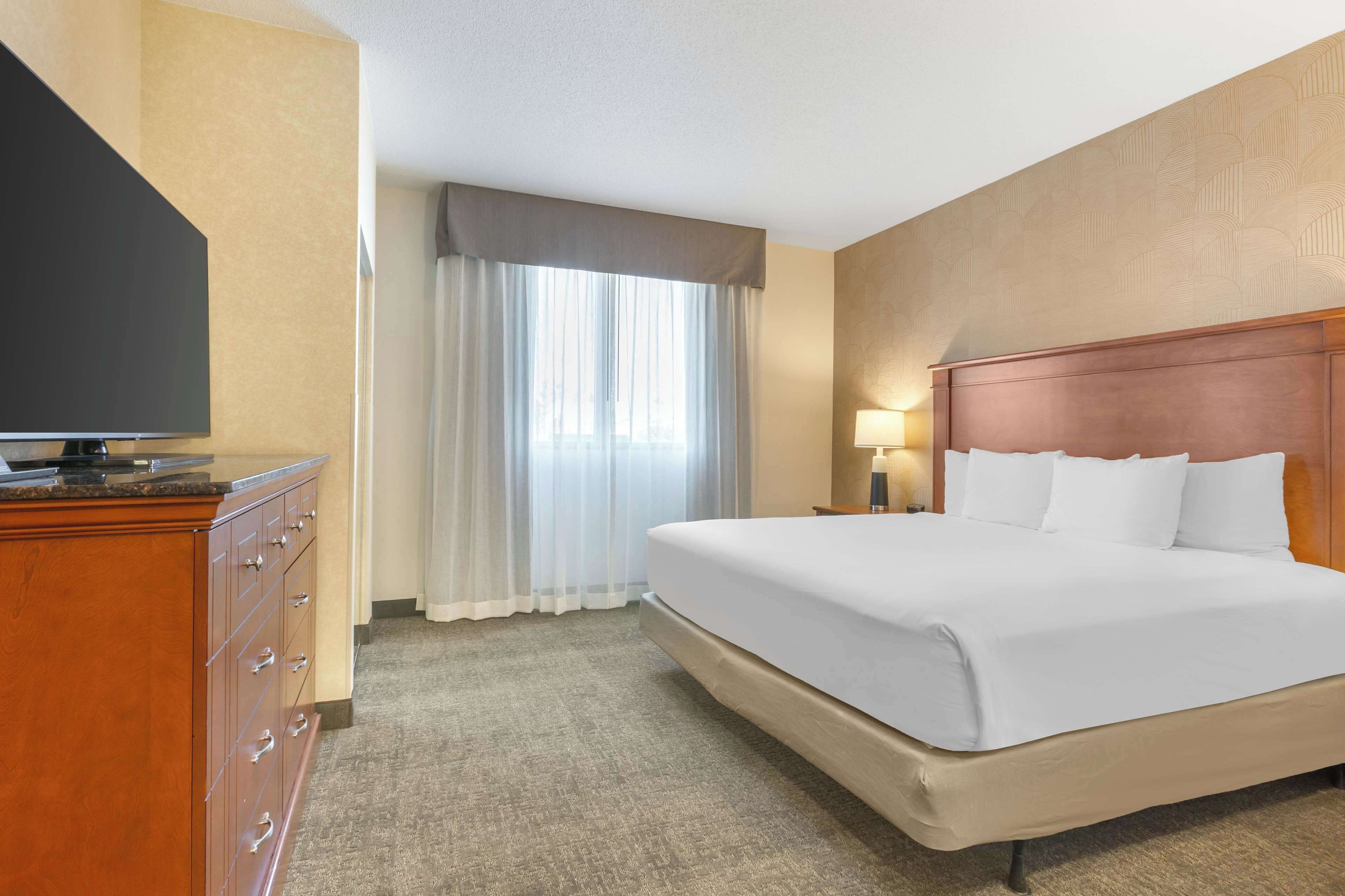 A hotel room with a large bed, a flat-screen TV on a wooden dresser, and a window with sheer curtains.