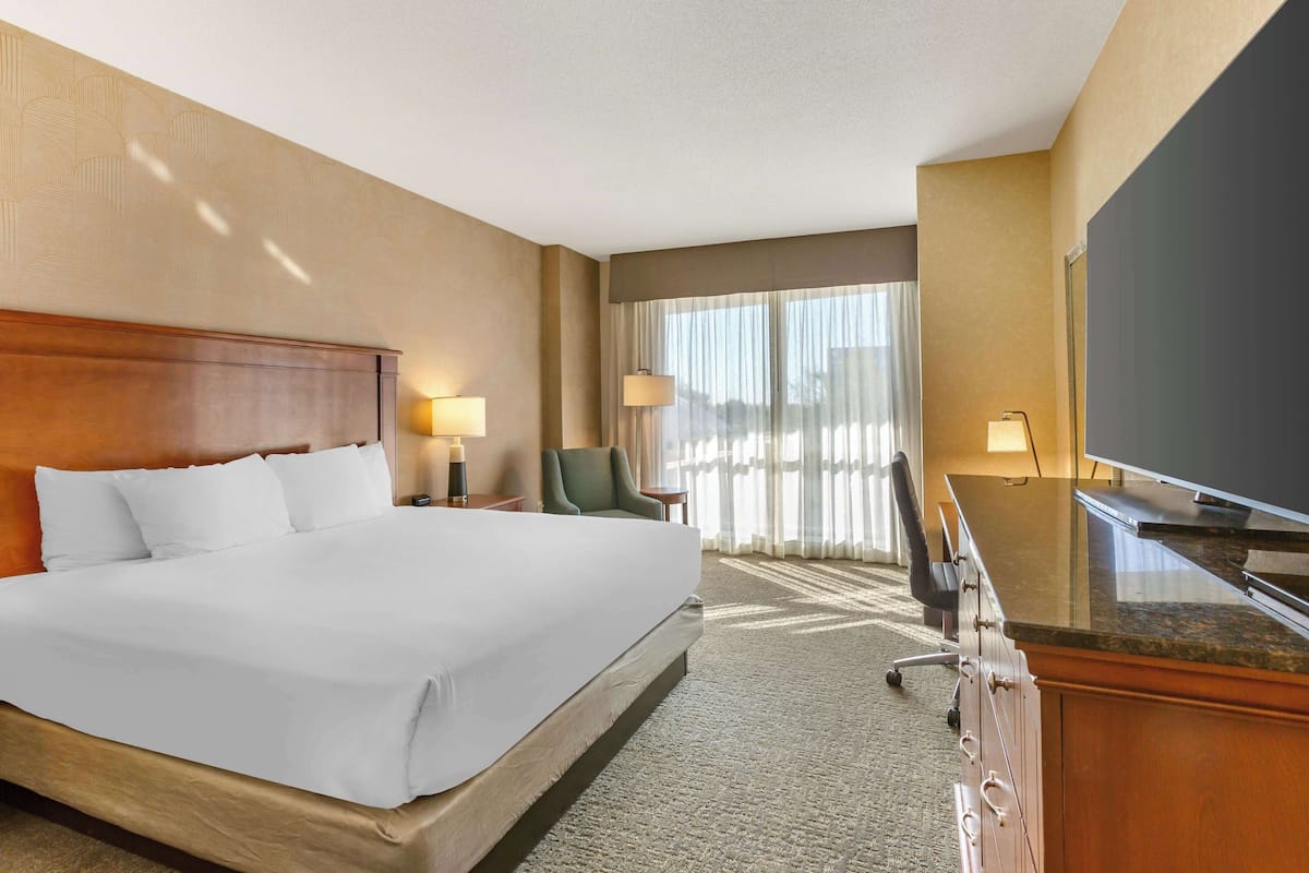 Habitación de hotel con una cama grande, un escritorio con televisor de pantalla plana, una silla, una lámpara y una ventana con cortinas traslúcidas.