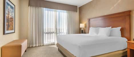 Quarto de hotel com uma cama grande, um banquinho, uma mesa de cabeceira e uma janela com cortinas.
