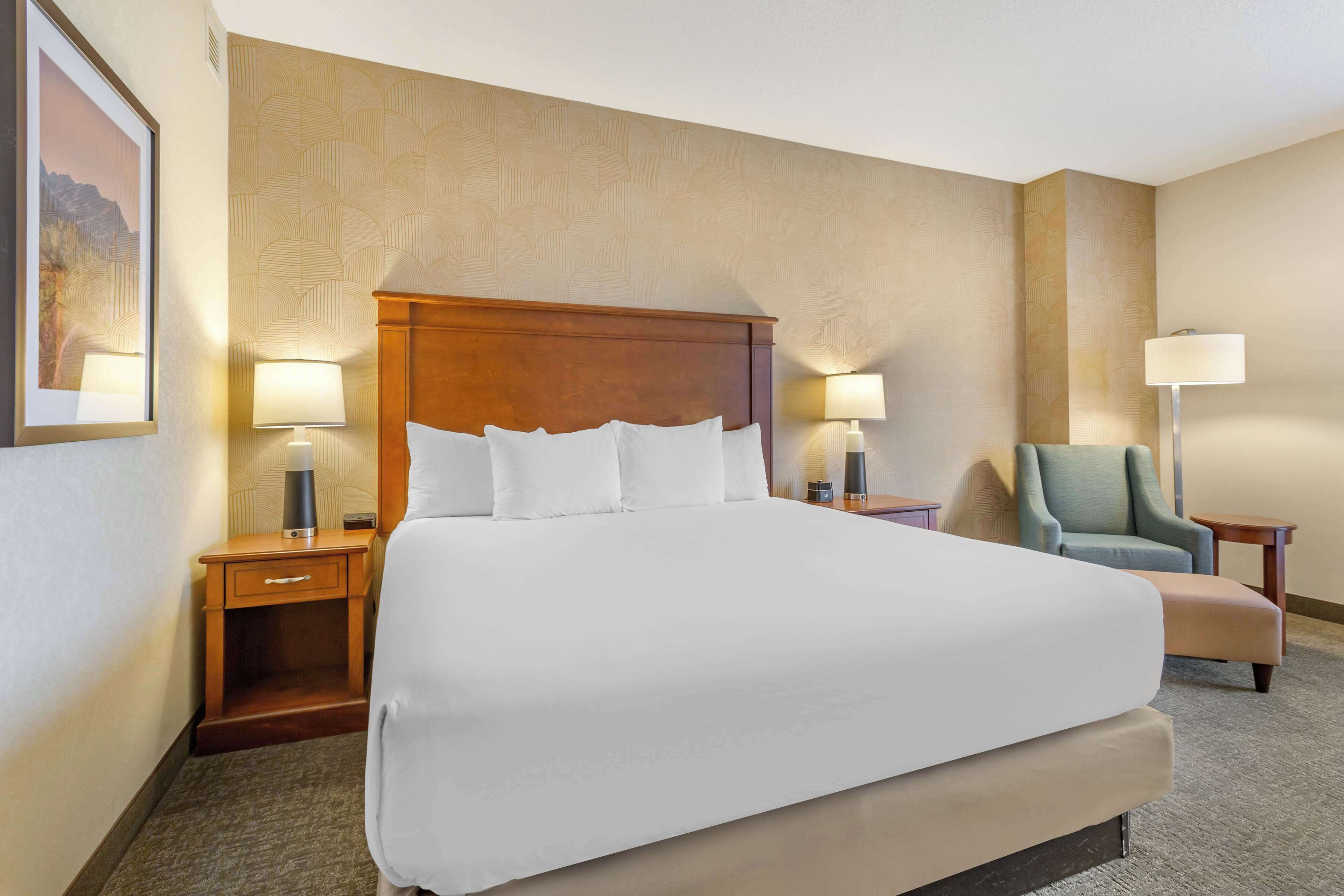 Habitación de hotel con una cama grande, dos mesitas de noche con lámparas, una silla e imagen enmarcada en la pared.