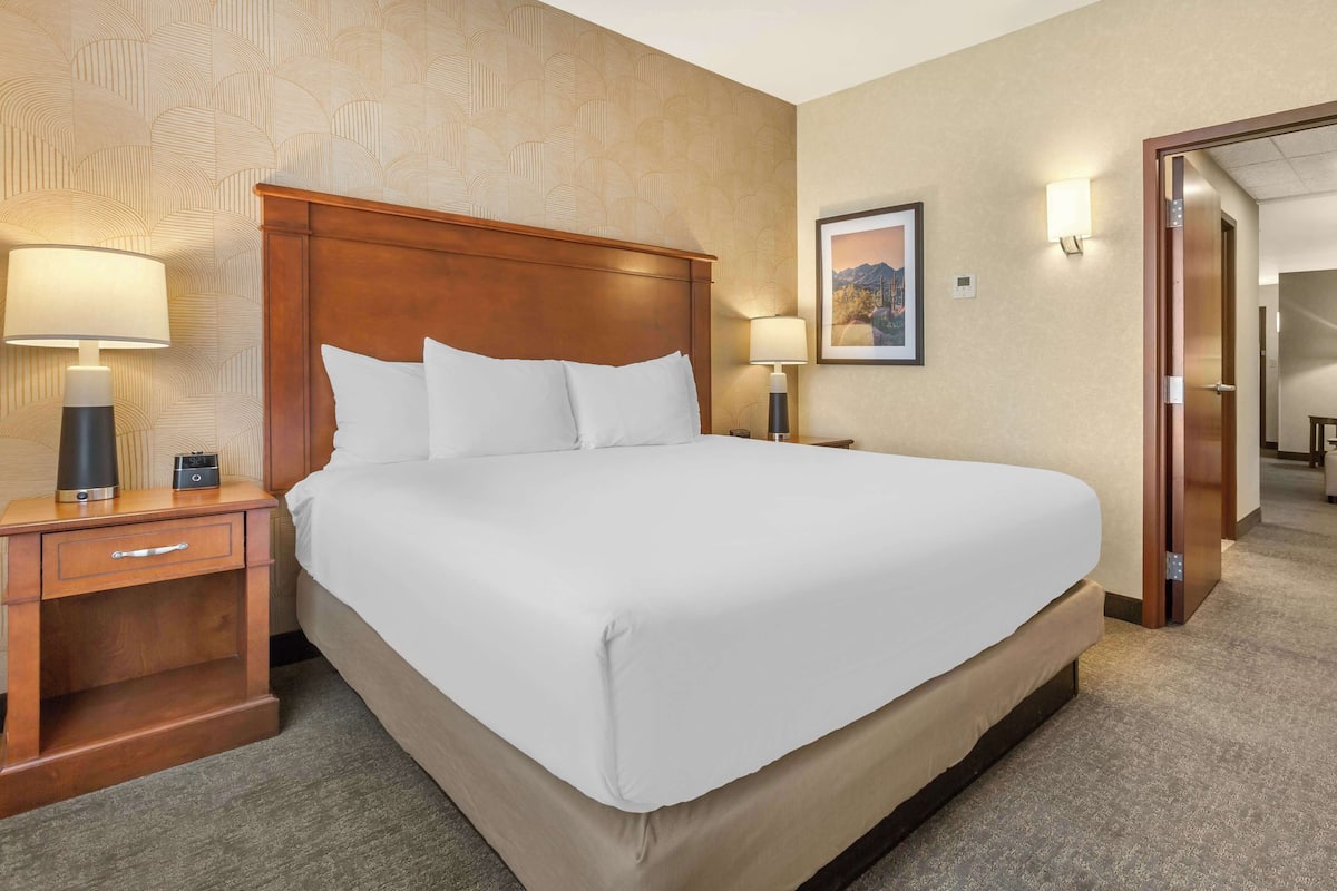 A hotel room with a large bed, wooden headboard, two bedside tables with lamps, a framed picture on the wall, and a carpeted floor.