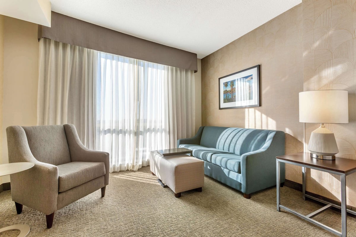 A hotel room with a beige armchair, a blue sofa, a small ottoman, a side table with a lamp, and a framed picture on the wall.