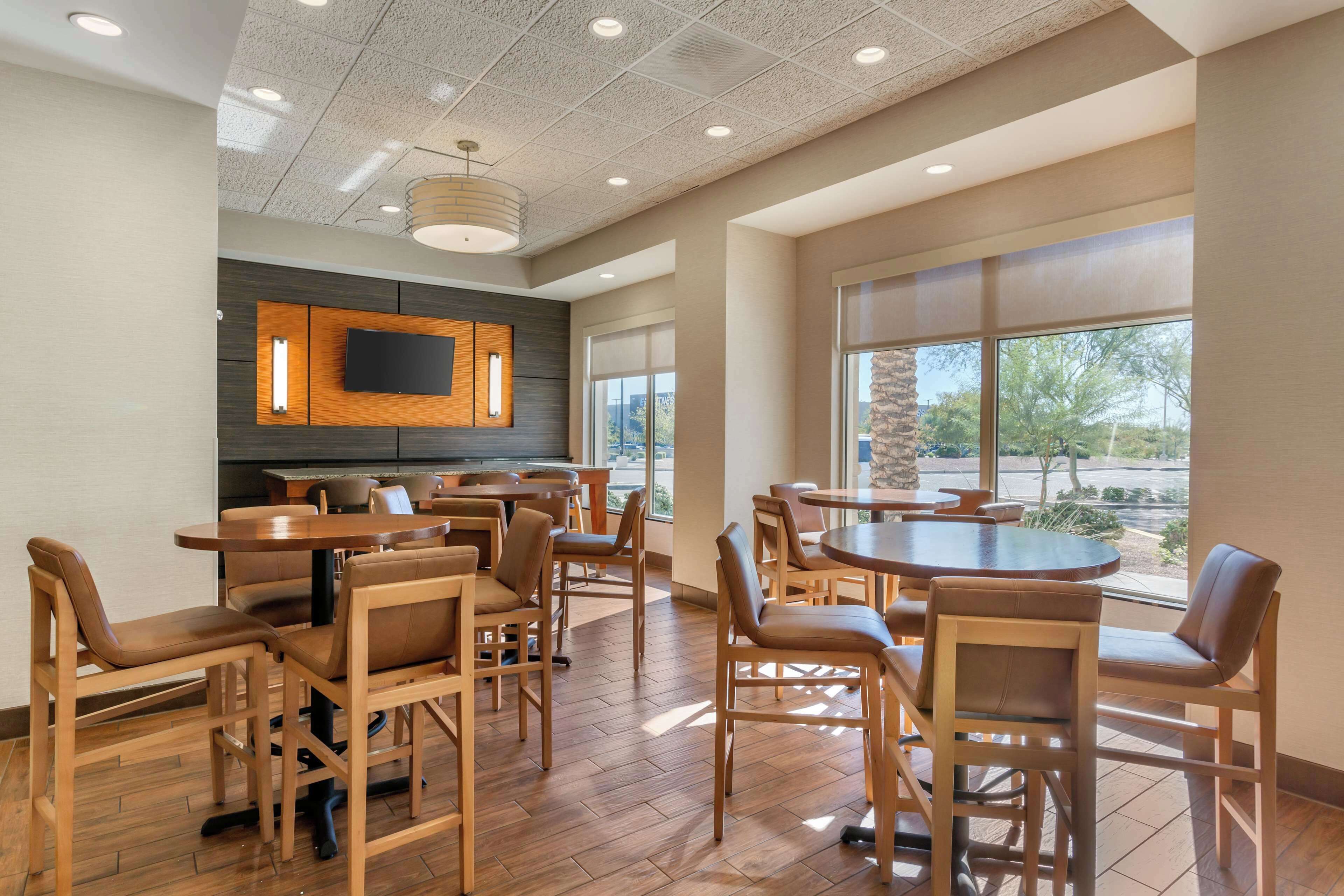 A restaurant with wooden tables and chairs, a flat-screen TV mounted on a wooden panel, and large windows offering a view of the outdoors.