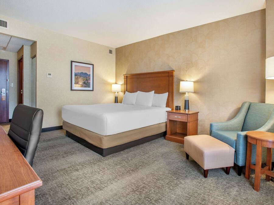 A hotel room with a large bed, a desk, a chair, a small table, and two lamps.